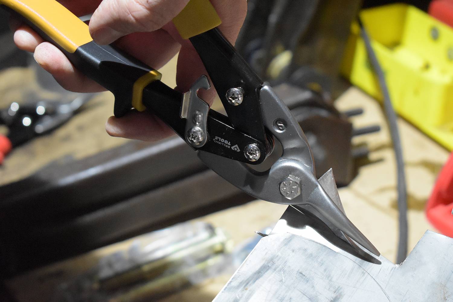 A large metal shears cuts through a sheet of metal on a workbench. Tools and accessories can be seen in the background.