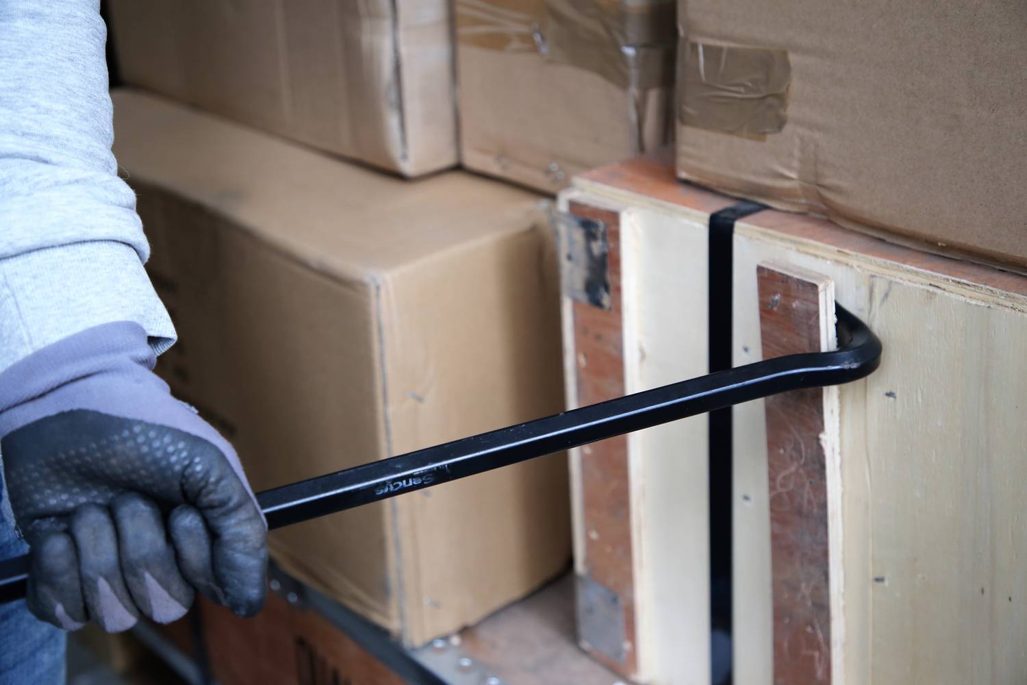 A person is levering open a wooden crate with a crowbar. The crate is made of wood, and other cardboard boxes are stacked around it.
