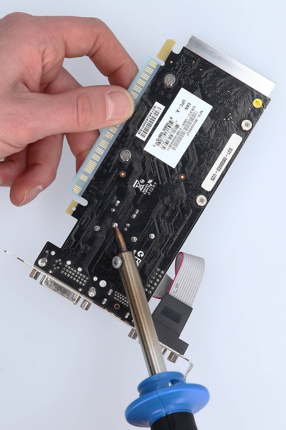 A hand holds a graphics card, while a soldering iron touches a pin on the circuit board, indicating repair work.