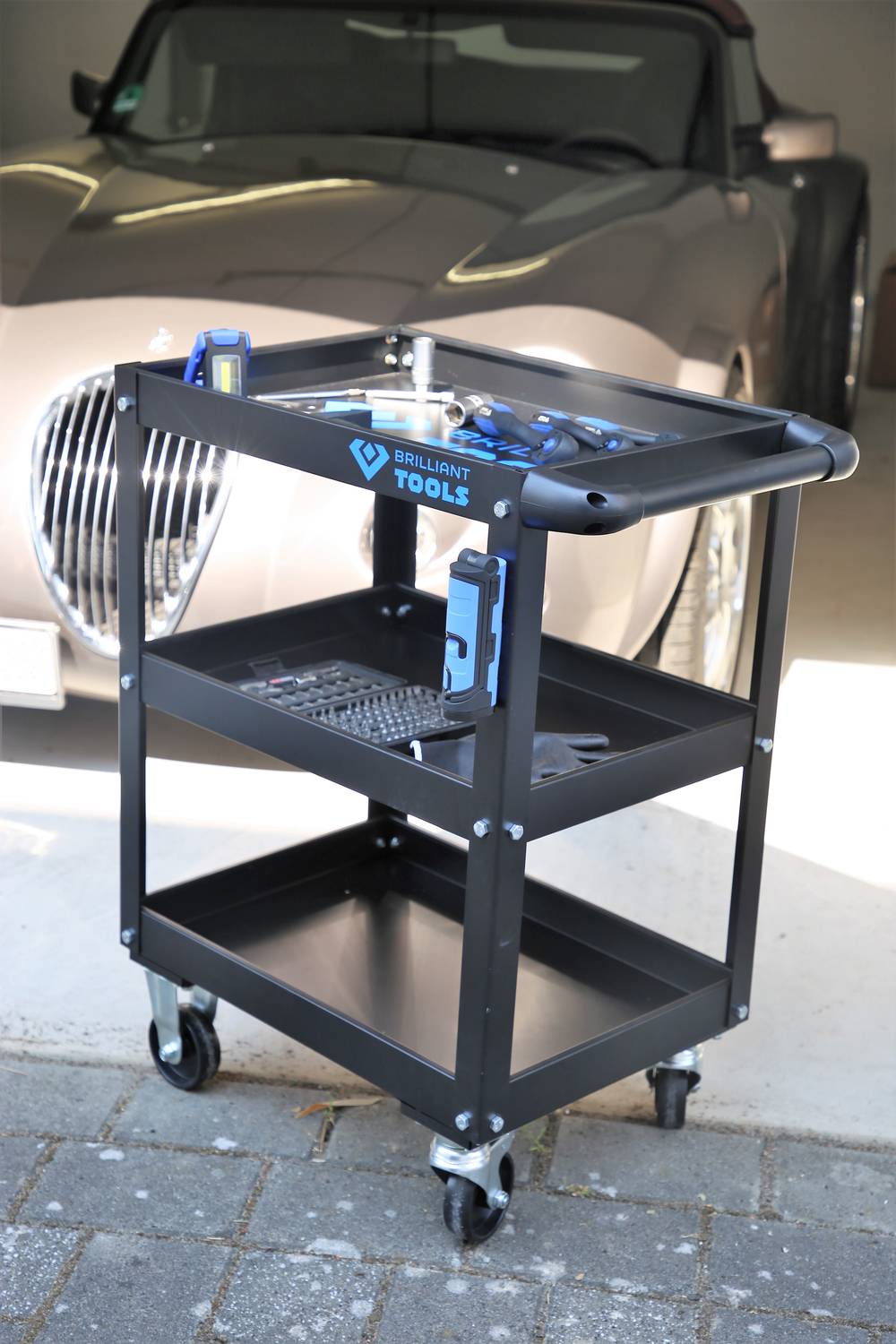 A black tool trolley with two shelves and various tools is standing in front of an elegant, gleaming car in a garage.