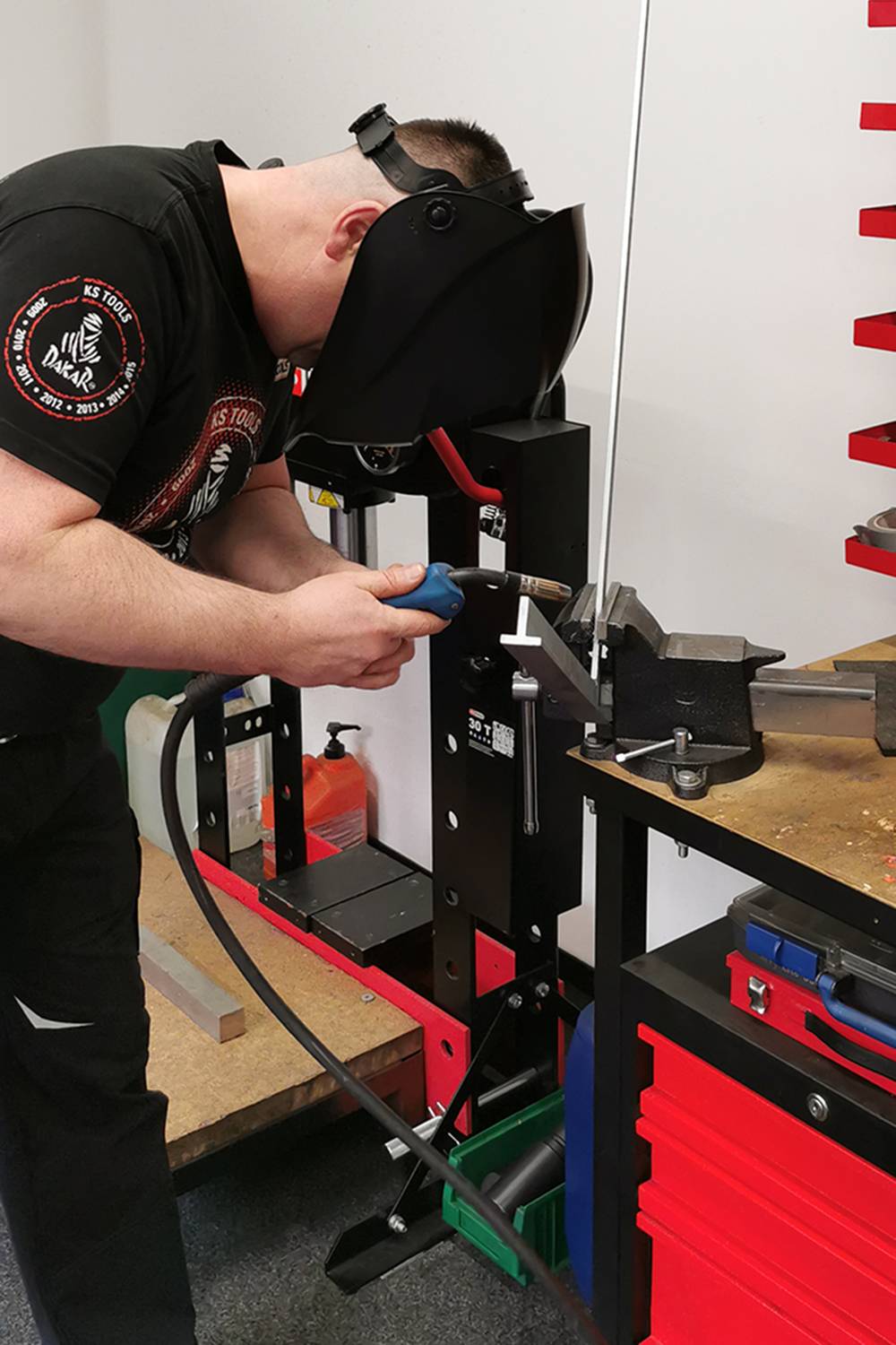 A man wearing a welding helmet is working on a metal piece in a workshop. He is using a welding machine next to a vice.