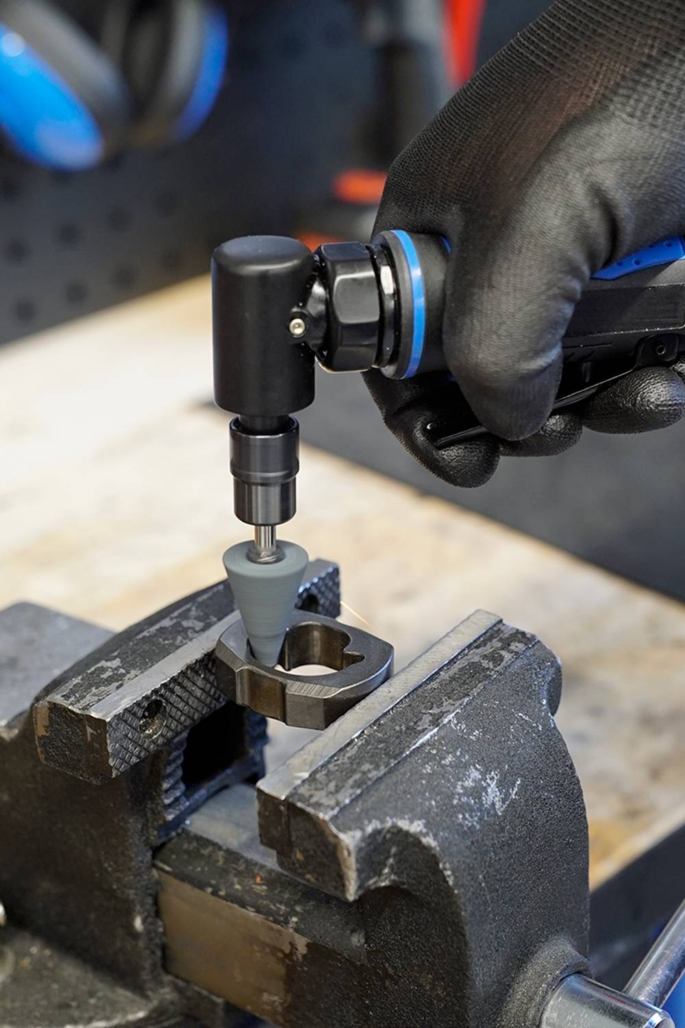 A person wearing a black glove is using an electric tool to machine a metal part in a vice.
