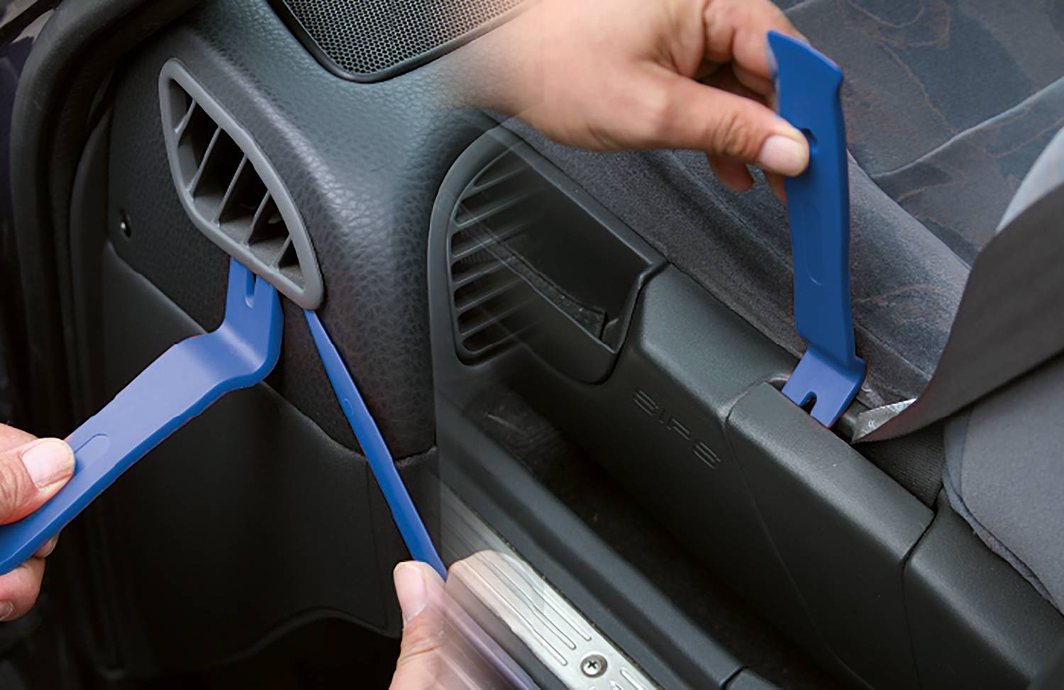 Two blue plastic tools are being used to safely remove vehicle trim panels. Hands are holding the tools near the ventilation and door trim.