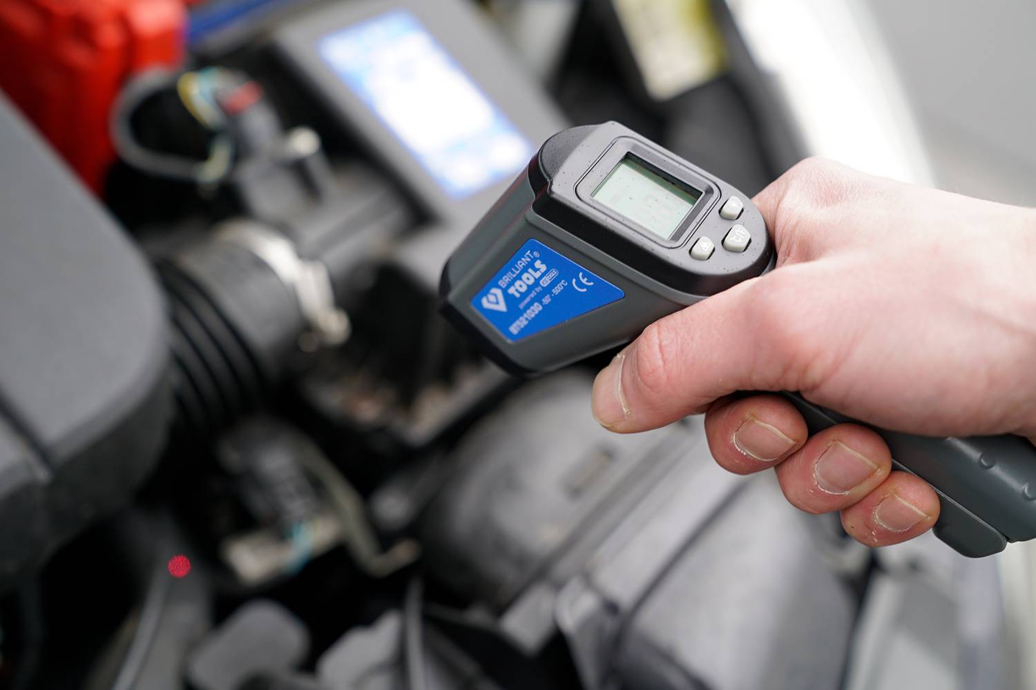 A hand is holding an infrared thermometer over a car part, measuring the engine temperature. The display shows a digital reading.