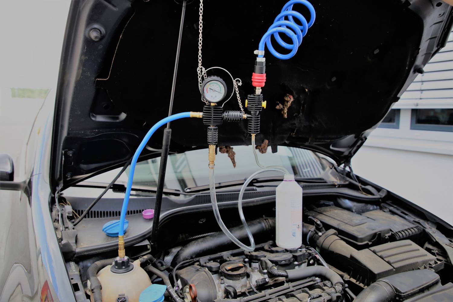 A car with an open bonnet, with a pressure gauge connected to it. A hose is connected to a liquid bottle.