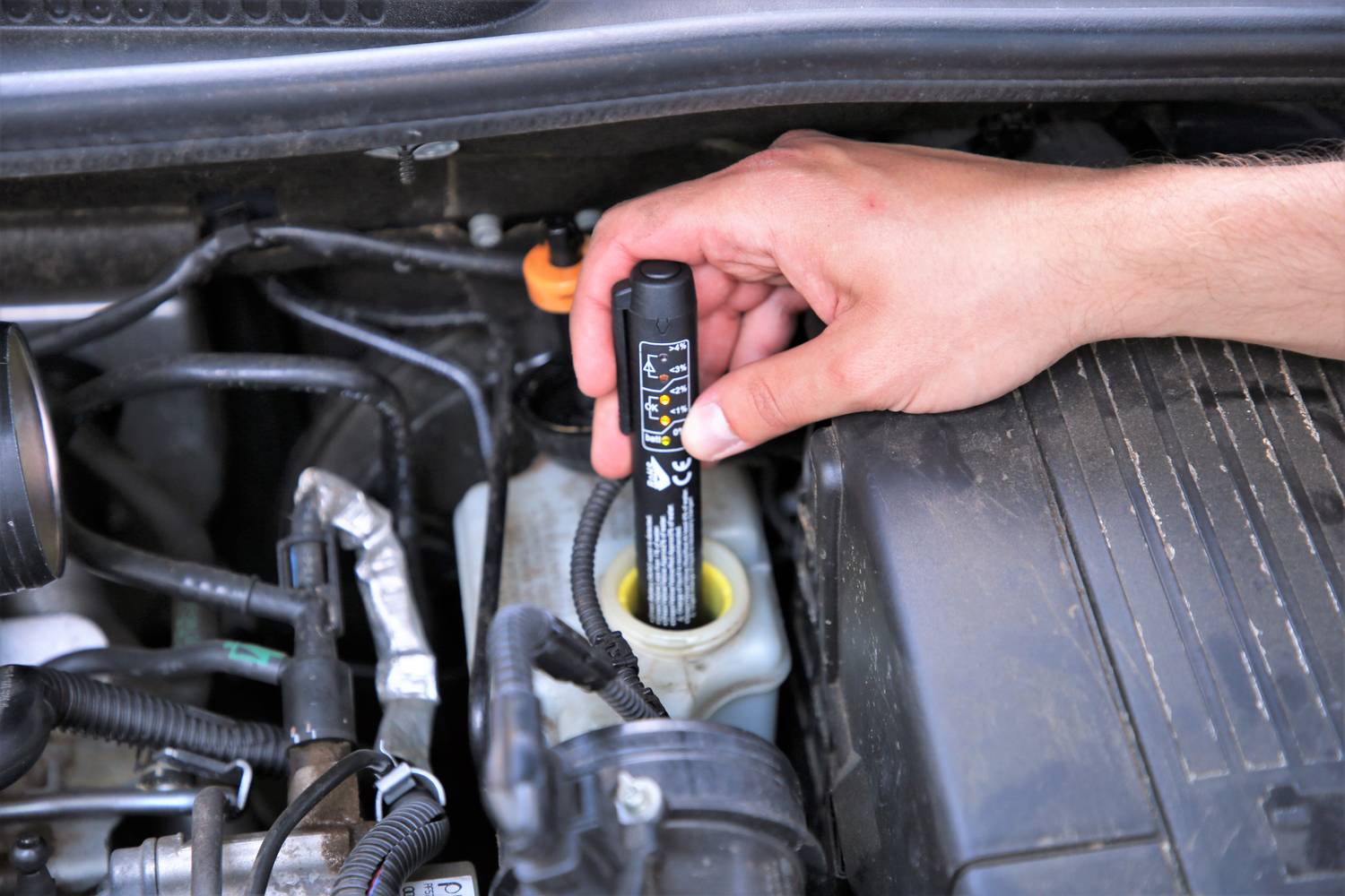 A hand is holding a brake fluid tester over the reservoir in the engine bay of a car to check the brake fluid.