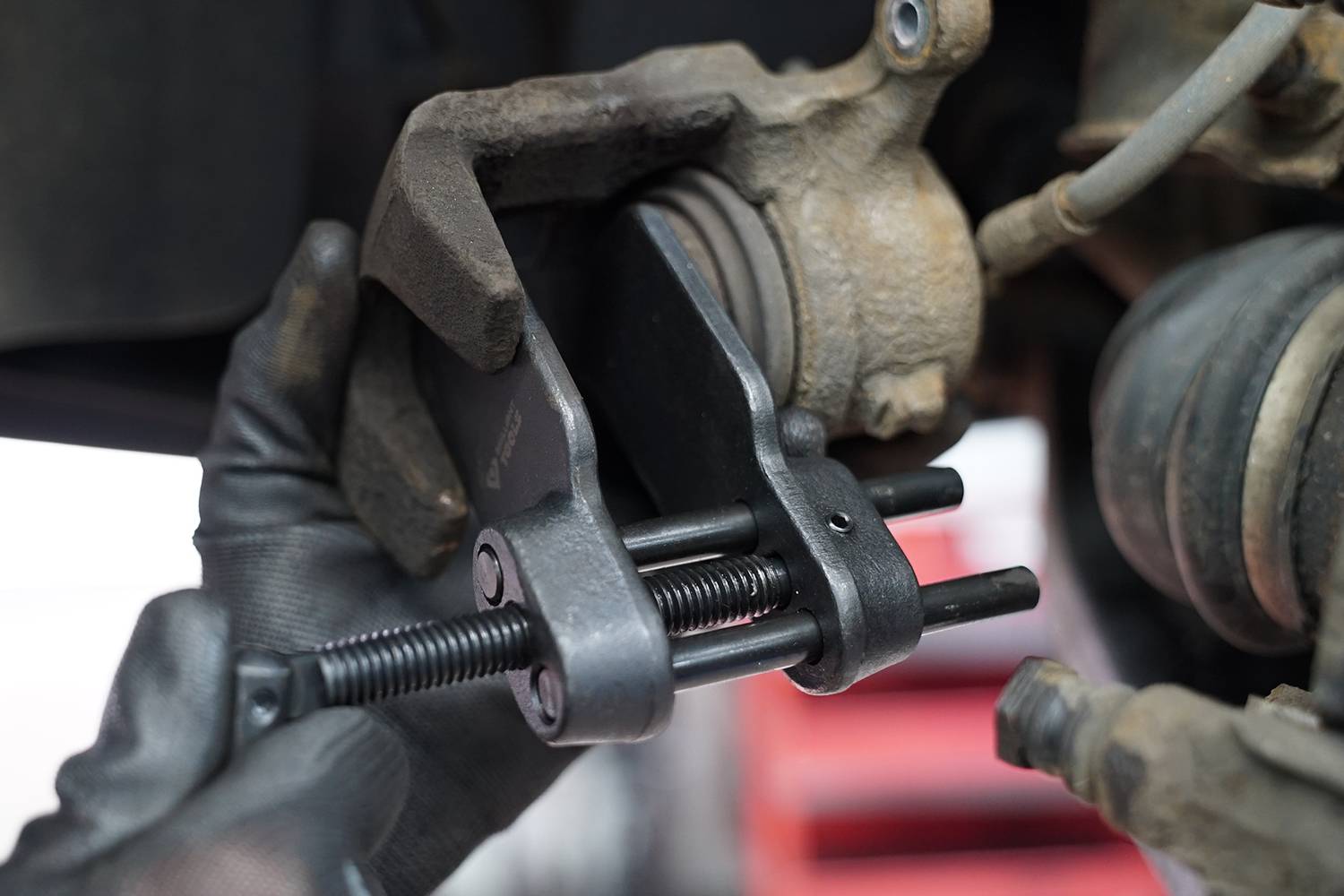 A person wearing gloves is using a tool to adjust a car's brake calliper. The tool is applying pressure to open the calliper.