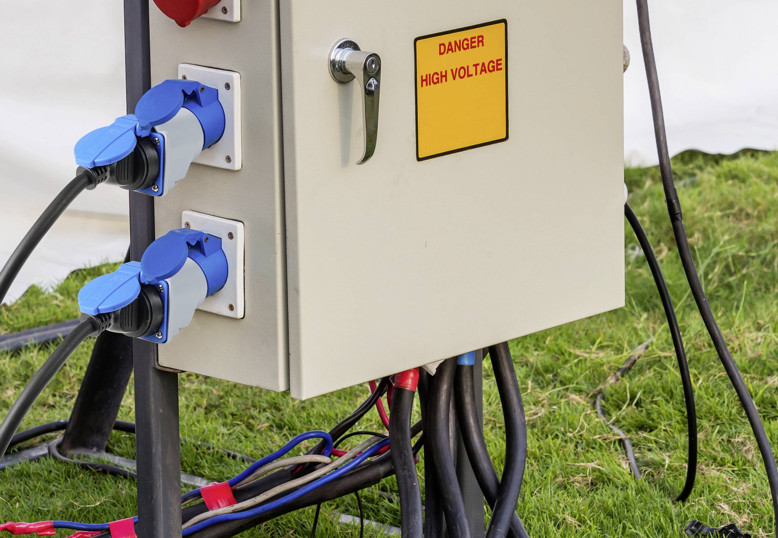 Electrical control cabinet with three blue power sockets and warning sign 'Danger: High Voltage', positioned on a meadow.