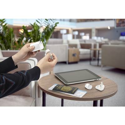 A person is holding a travel adapter in a waiting area; a tablet, passport, and boarding pass are lying on the table.