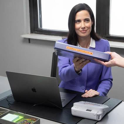 A woman in a suit is sitting at a desk with a laptop and printer. She is handing over a folded brochure with orange text.