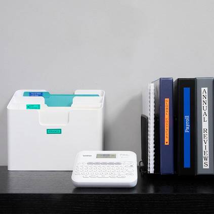 On a table are folders and a label printer. To the left are sorted files, and to the right are books with labels such as 'Payroll' and 'Annual Reviews'.