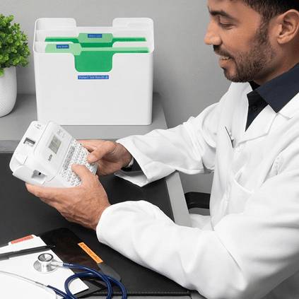 A man in a white lab coat sits at a table and examines a device. A green storage basket is in the background.