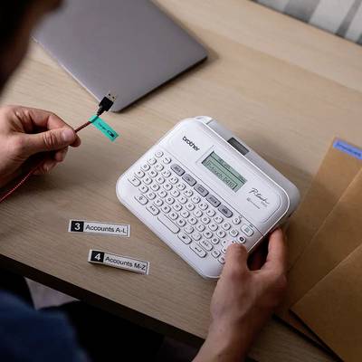 A person is using a label printer to print labels. A laptop and envelopes are lying on the table.