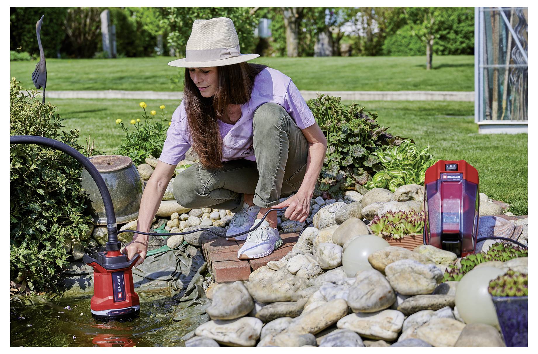 Woman in a sunhat crouches by a garden pond, adjusting a red water pump or fountain among rocks and plants.