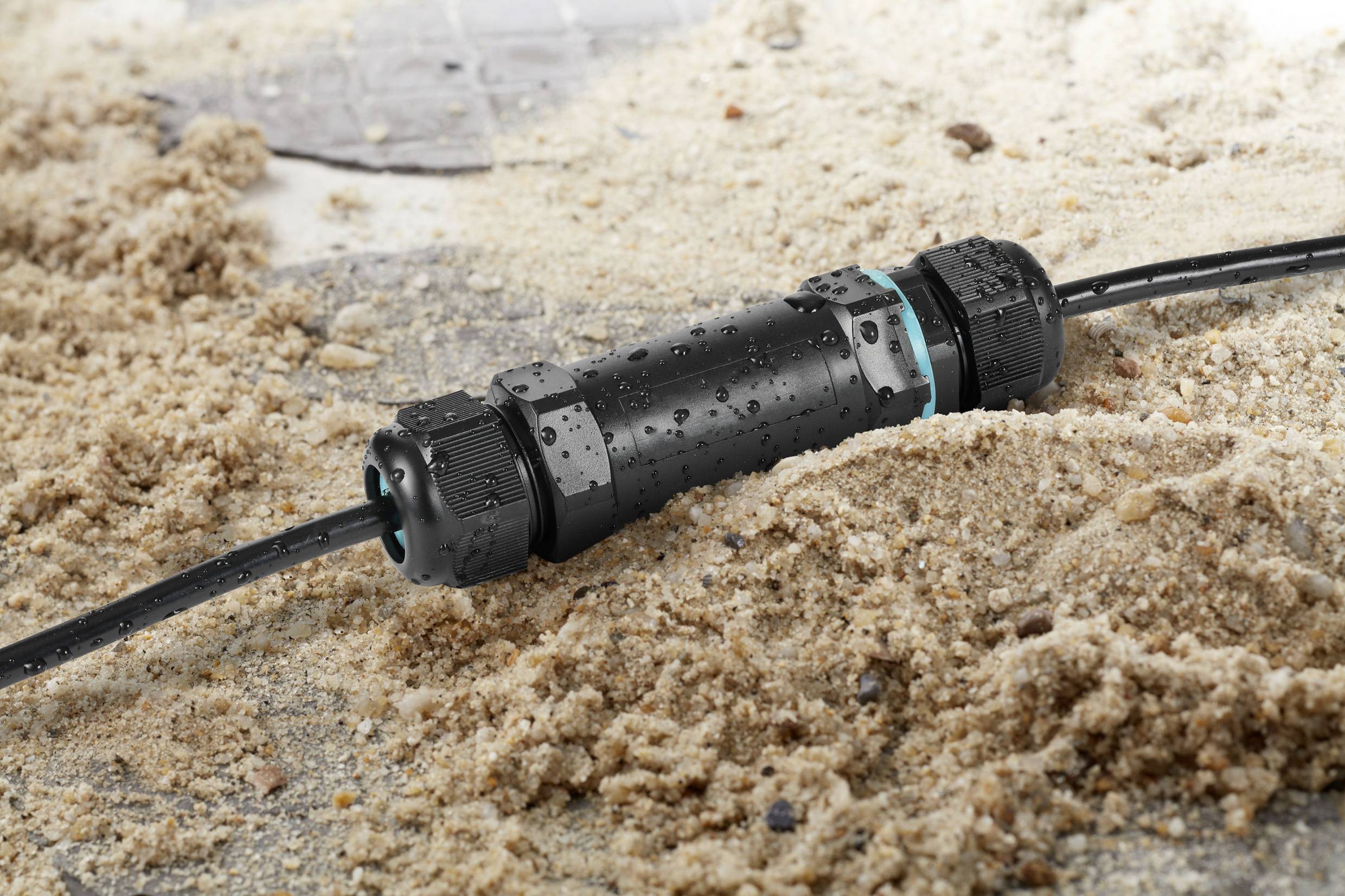 A waterproof cable connector lies in the sand. The cable is connected and demonstrates protection against moisture and sand in an outdoor environment.