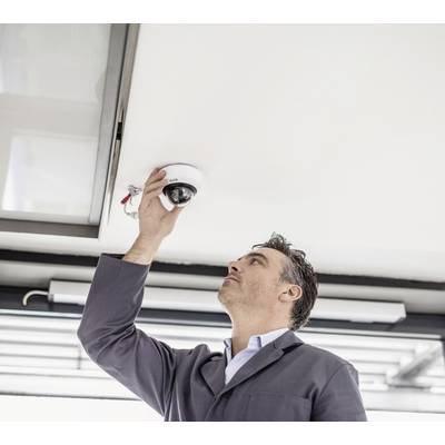 A man in workwear is examining or installing a surveillance camera on the ceiling in a modern building.