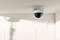Security camera mounted on the ceiling of a bright room, pointing towards a corner with white brick walls.