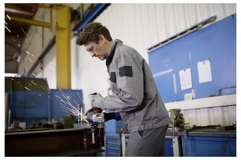 A person in protective gear uses a grinder, creating sparks, in an industrial workshop with blue and white walls in the background.