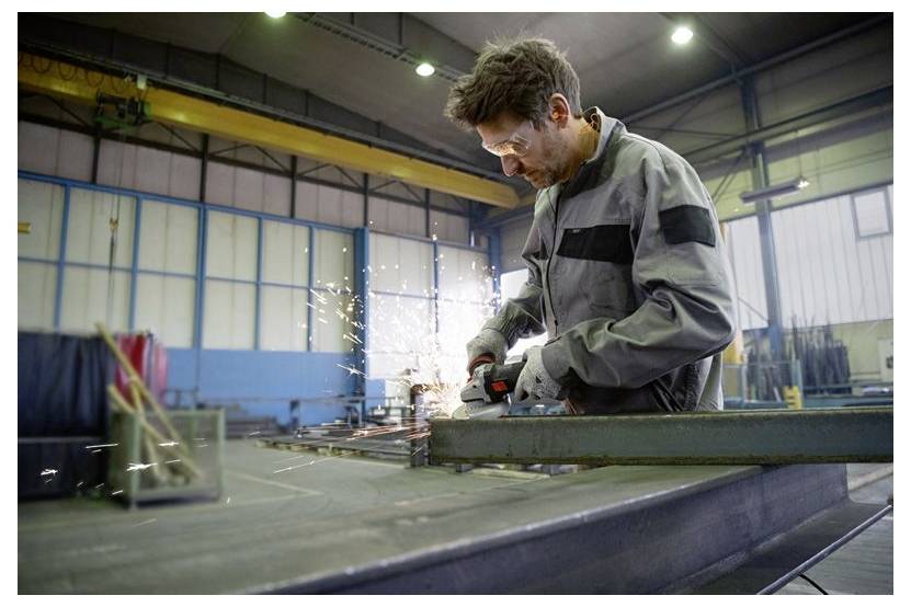 A person in a workshop using a grinder on a metal beam, with sparks flying in a large industrial setting.