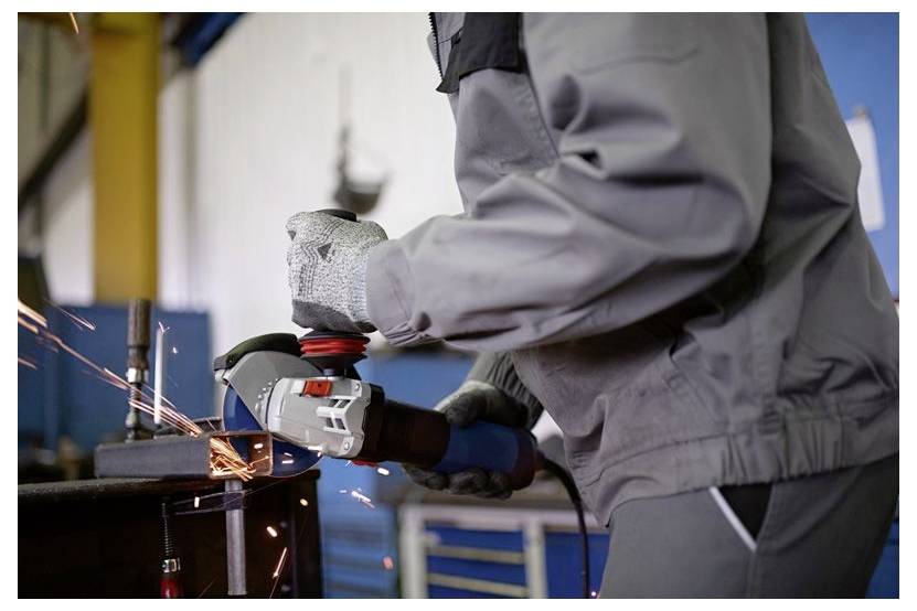 A person wearing protective clothing and gloves uses an angle grinder on metal, creating sparks in an industrial workshop setting.
