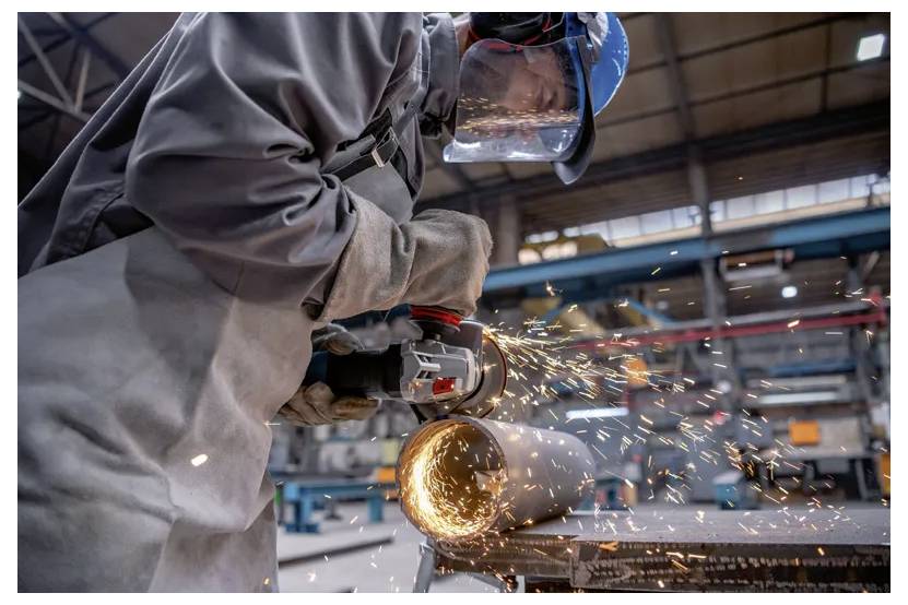 A worker wearing protective gear uses an angle grinder to cut a metal pipe, producing sparks in an industrial workshop setting.