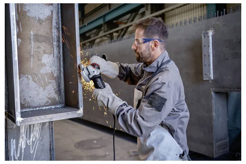 A worker in protective gear uses an angle grinder to cut metal, with sparks flying, in an industrial setting.