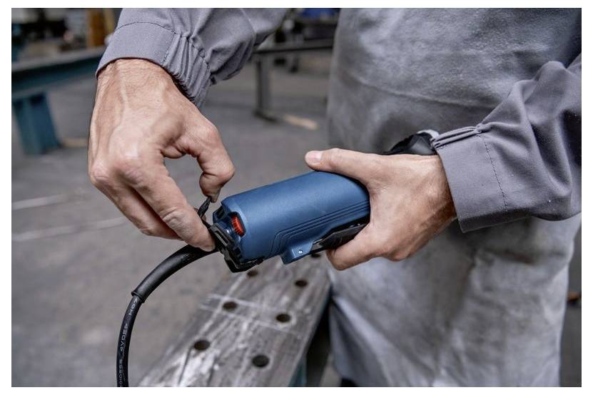 A person in work attire uses a blue power tool in a workshop, focusing on adjusting the settings or attaching a component.