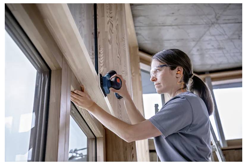 A person using a power sander to smooth a wooden window frame. They are wearing safety glasses and ear protection in a well-lit room.