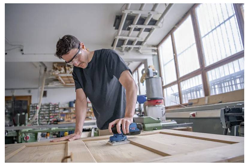 A person wearing safety glasses sands a wooden surface in a workshop. Various tools and equipment are visible in the background.