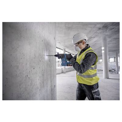 A construction worker wearing a hard hat and safety vest uses a power drill on a concrete wall in an unfinished building interior.