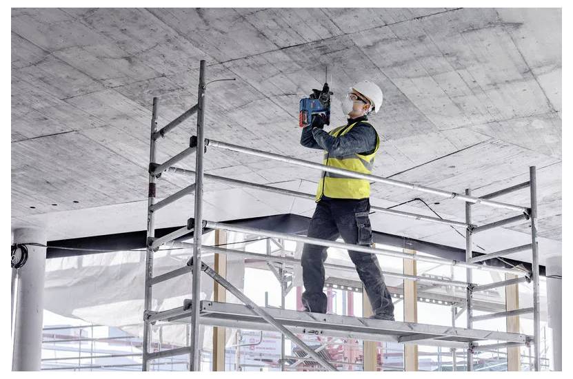 A construction worker in a safety helmet and vest uses a power tool on a ceiling while standing on a scaffolding platform in a building site.