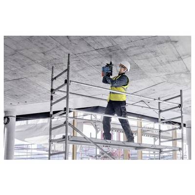 A construction worker in a safety helmet and vest uses a power tool on a ceiling while standing on a scaffolding platform in a building site.