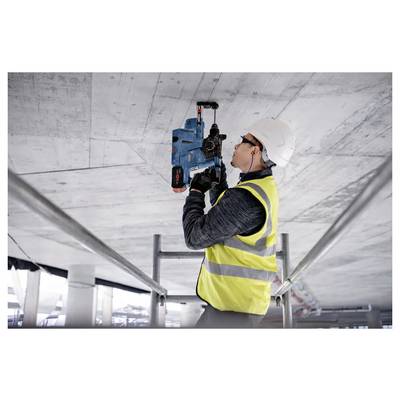 A construction worker in safety gear uses a power tool to drill into a concrete ceiling on a scaffolding, in an unfinished building.