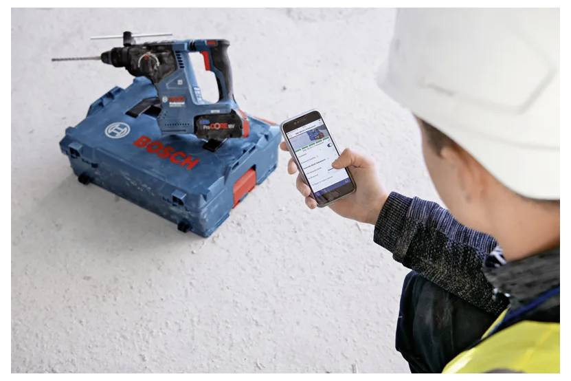 A person using a smartphone to control or monitor a Bosch power tool placed on the floor. The tool is in a blue carrying case.