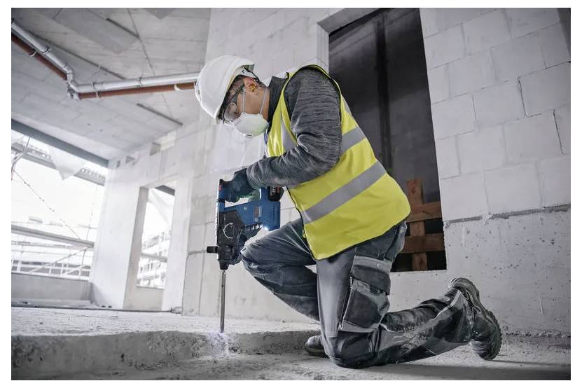 A construction worker wearing a safety vest, helmet, and mask kneels while using a drill in a partially constructed building.