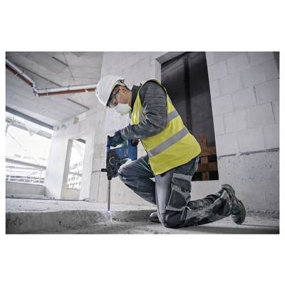 A construction worker wearing a safety vest, helmet, and mask kneels while using a drill in a partially constructed building.