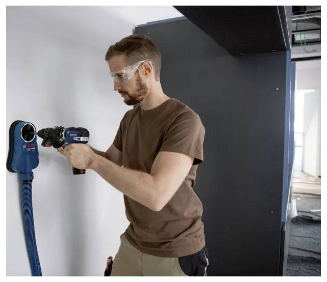 'A person wearing safety goggles uses a power drill with dust extraction on a white wall.'