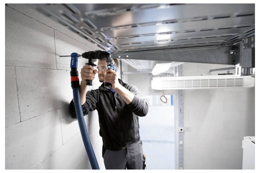 A person installing a ventilation duct in a concrete-walled room, using a tool to secure blue tubing under a metal frame near the ceiling.
