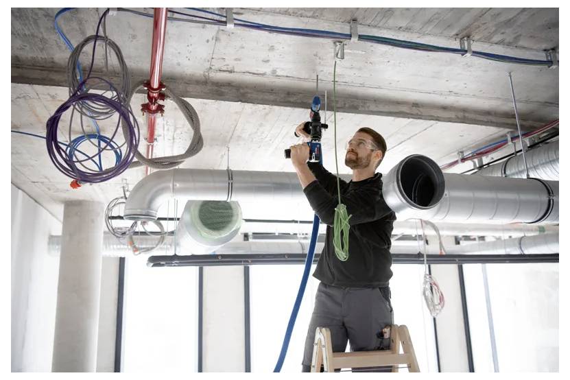Electrician on a ladder installing wiring on a ceiling in an industrial space, surrounded by exposed pipes and ducts.