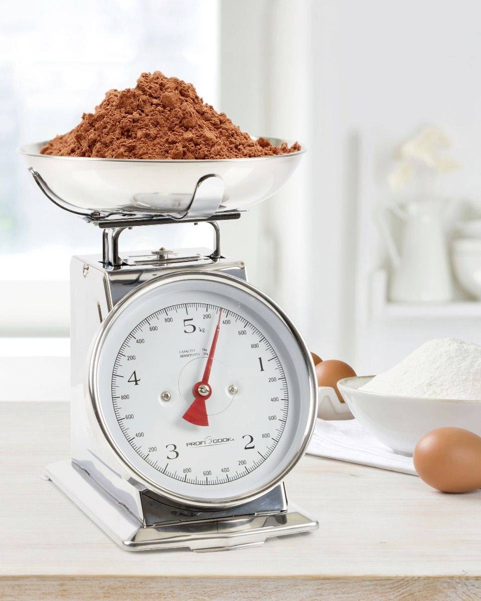 A kitchen scales with a bowl filled with cocoa powder, with eggs and a bowl of flour lying beside it on a wooden table.