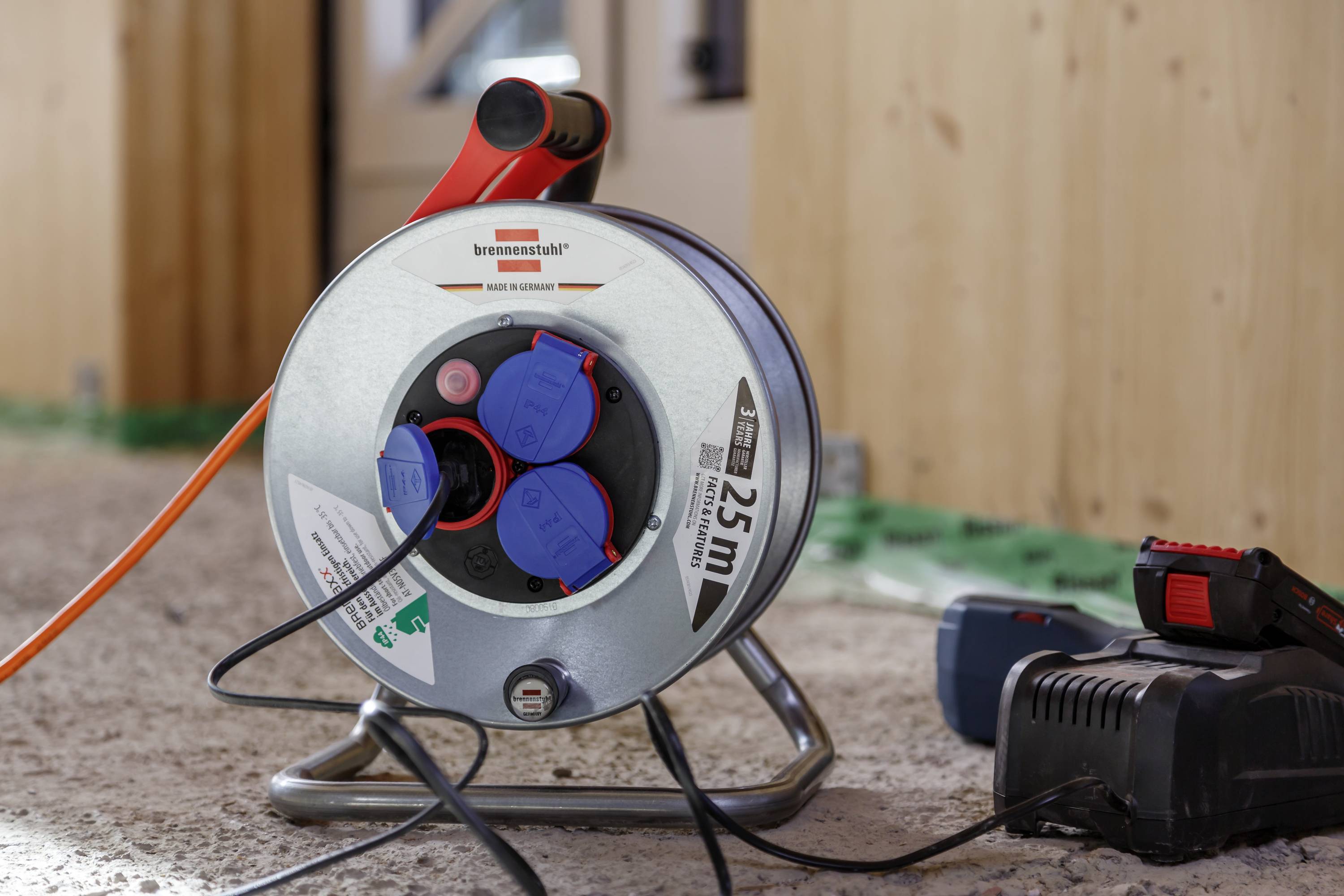 A cable reel with three blue plug sockets and a 25m label is standing on a floor, with a charger connected.