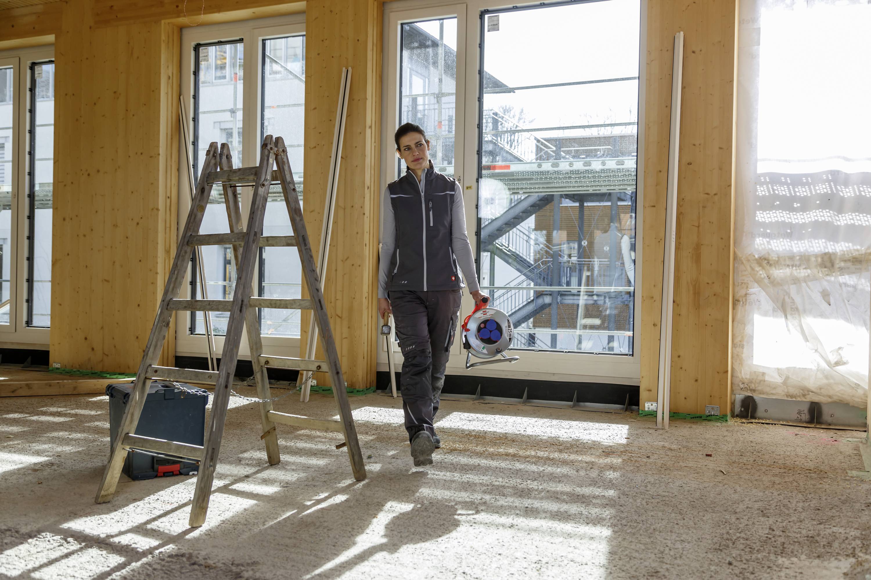 A person is standing in a partially completed room with wooden panelling and large windows, holding a cable drum. A ladder is standing next to them.