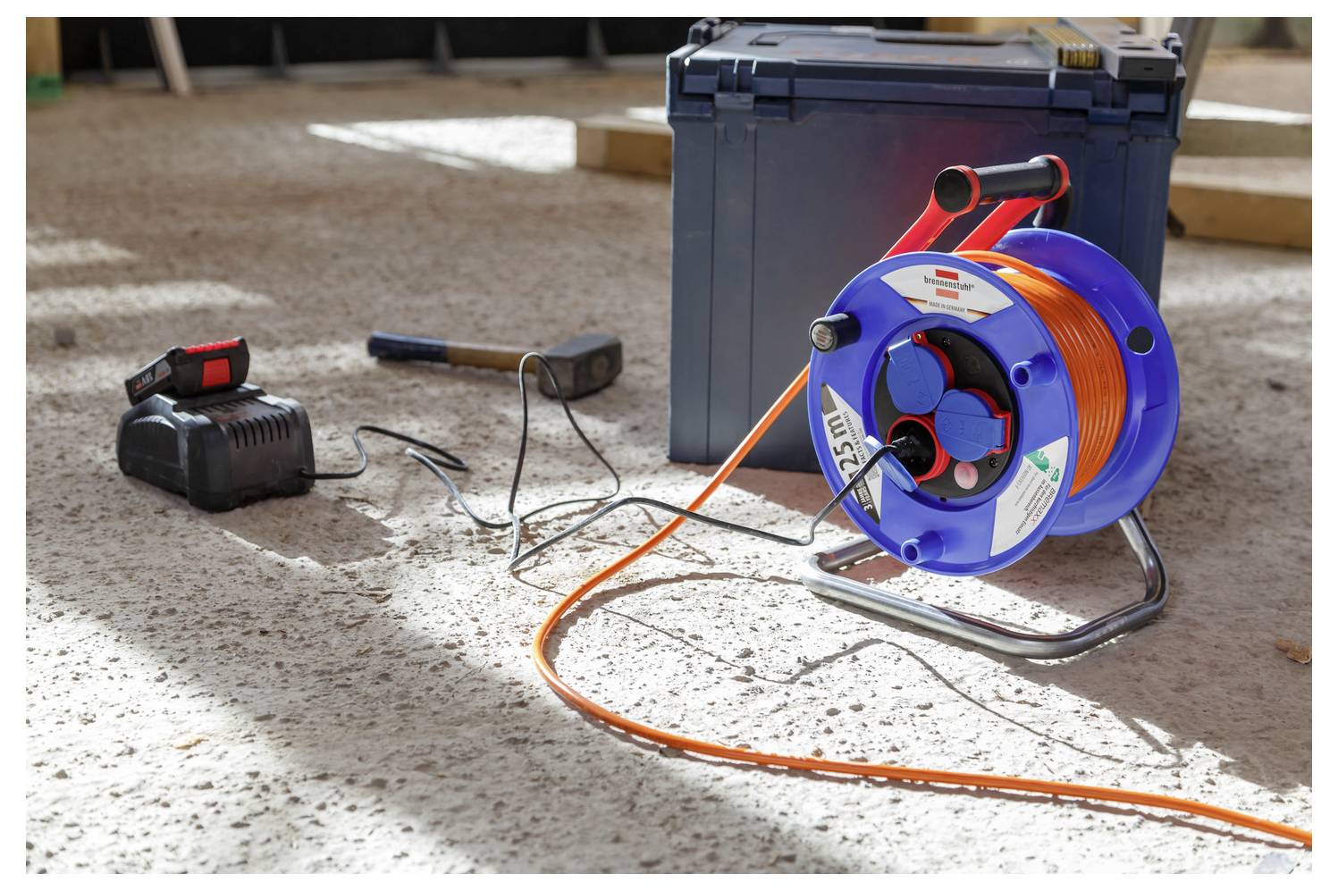 Portable extension cord reel with orange cable, battery charger, and toolbox on a concrete floor at a work site.