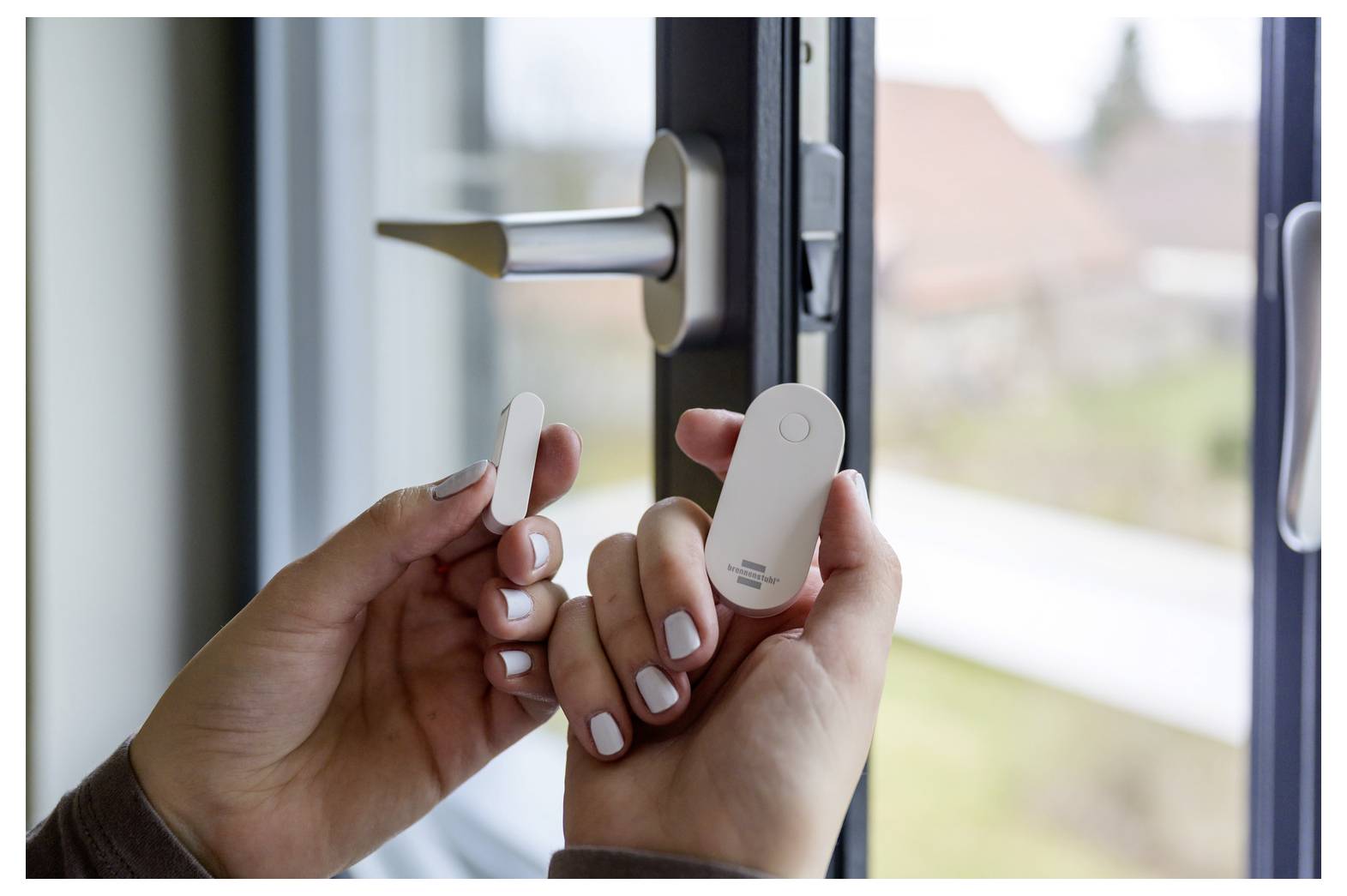 Hands holding a wireless door/window sensor near an open window latch, suggesting home security installation.