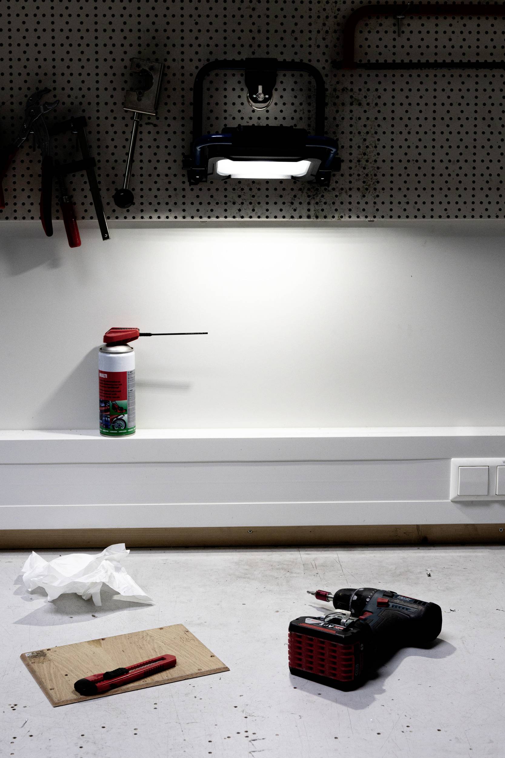Workbench in workshop with lamp. On the workbench lies a drill, a craft knife and a wooden board. Tools are hanging on the wall.