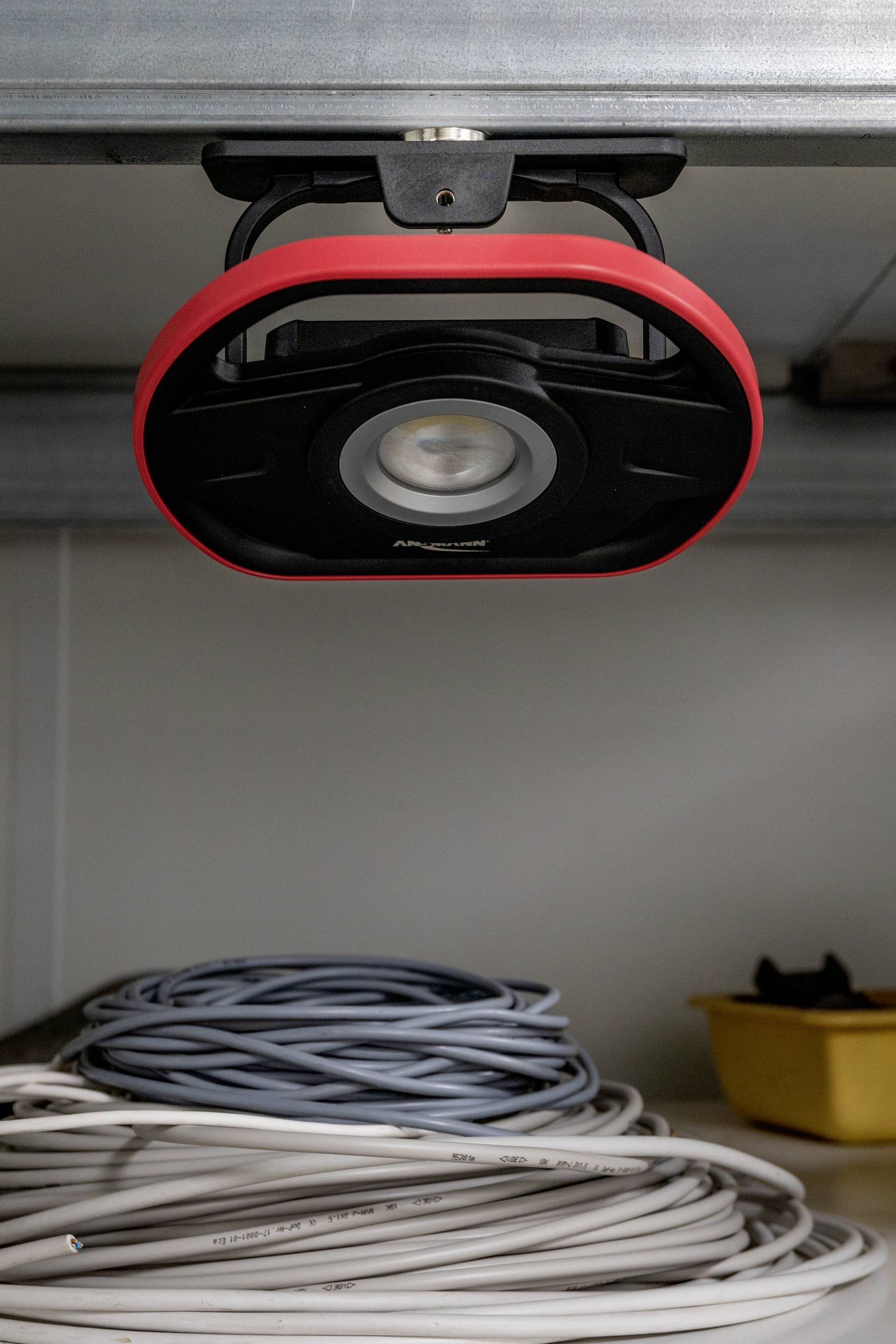 Lamp with a red frame mounted on the ceiling; in front of it, coils of grey and white cables; yellow container in the background.