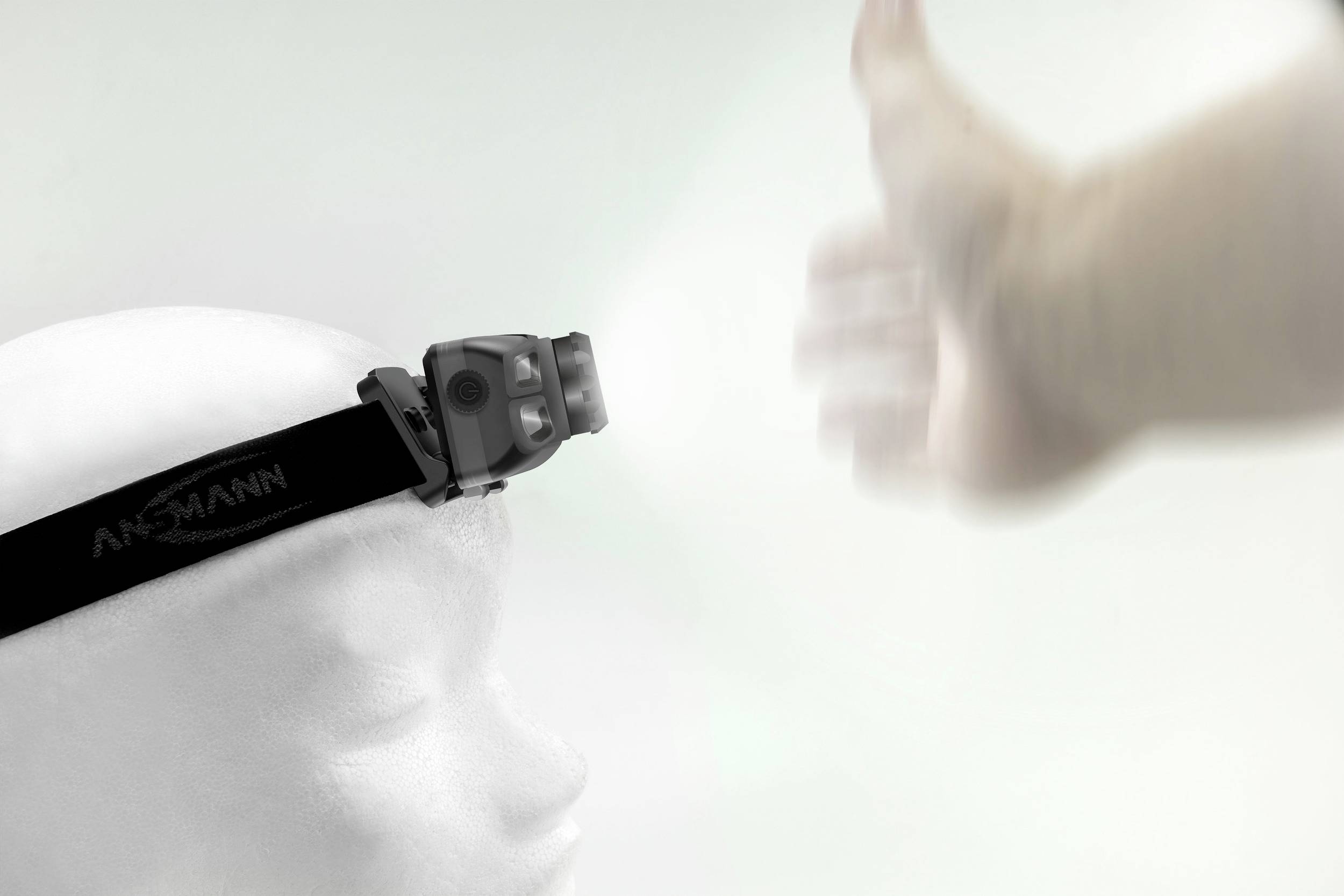 A white mannequin head is wearing a head torch that is switched on. A hand is visible moving in front of the light.