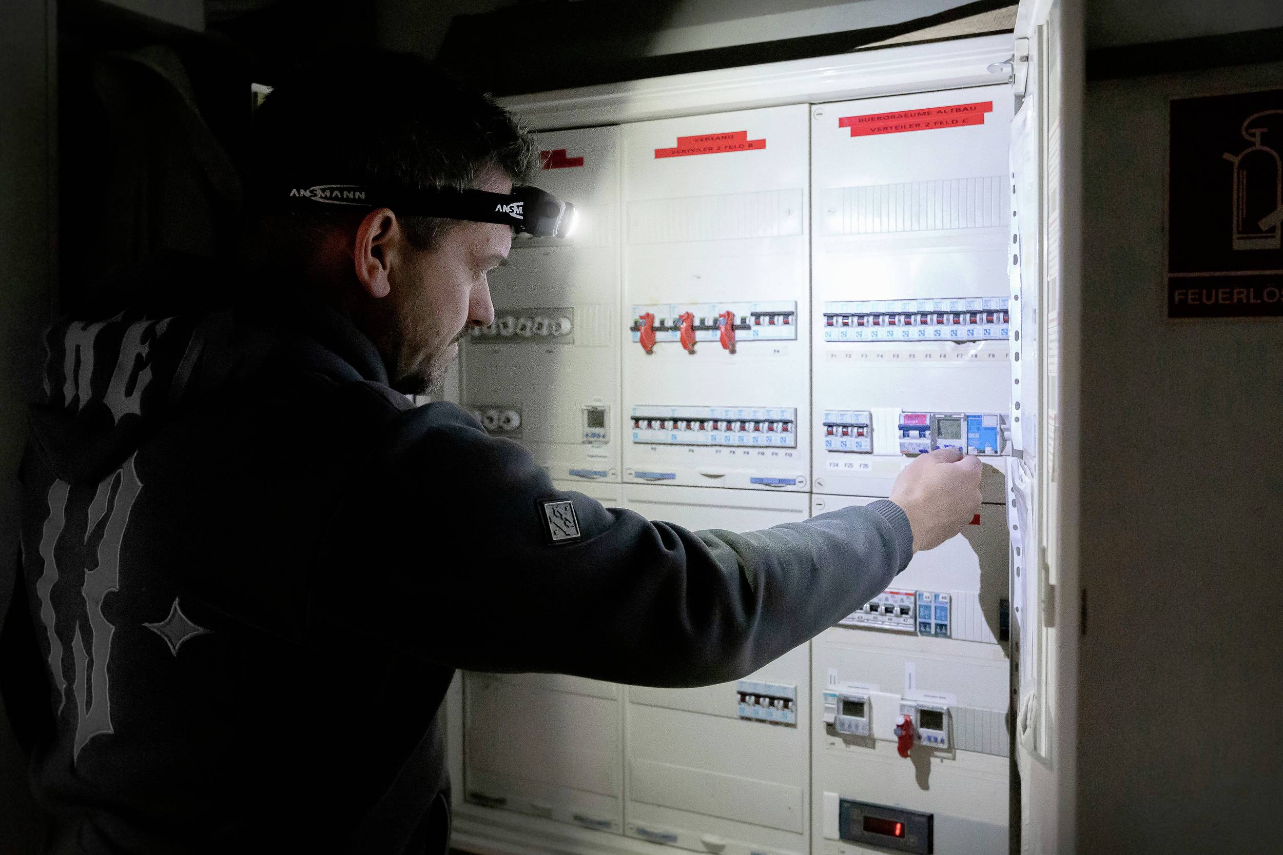 A man wearing a head torch is working on an open electrical cabinet, checking the fuses in a dimly lit room.