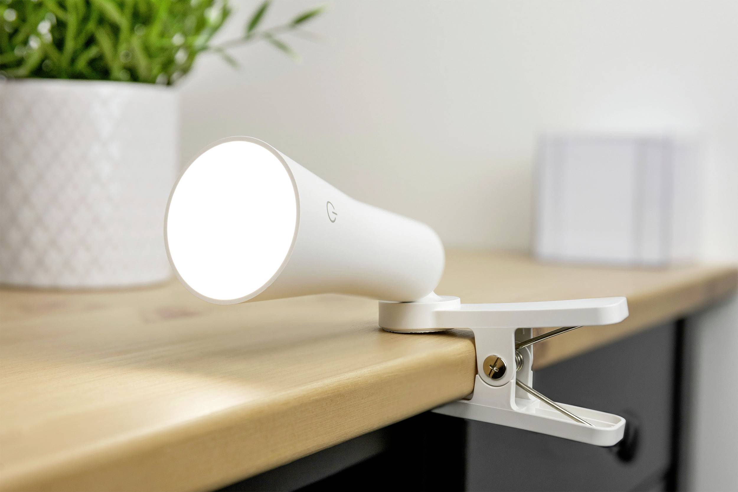 White LED clip-on lamp, fastened to the edge of a table, shines brightly. A plant and a cube-shaped object are blurred in the background.
