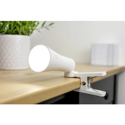 White LED clip-on lamp, fastened to the edge of a table, shines brightly. A plant and a cube-shaped object are blurred in the background.